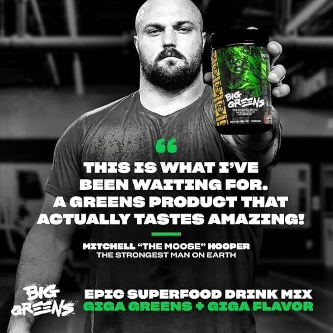 A man in a gym setting holds up a container of Big Greens Epic Superfood Drink Mix Giga Greens + Giga Flavor. The image features a black-and-white photo of the man with green and white text overlays promoting the product.