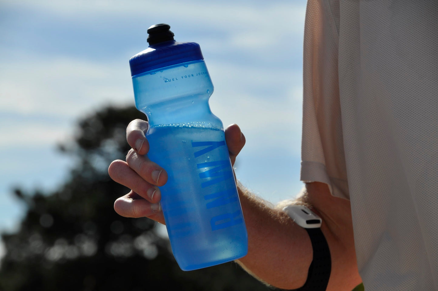 Person holding a blue water bottle with a brand logo against a blurred outdoor background