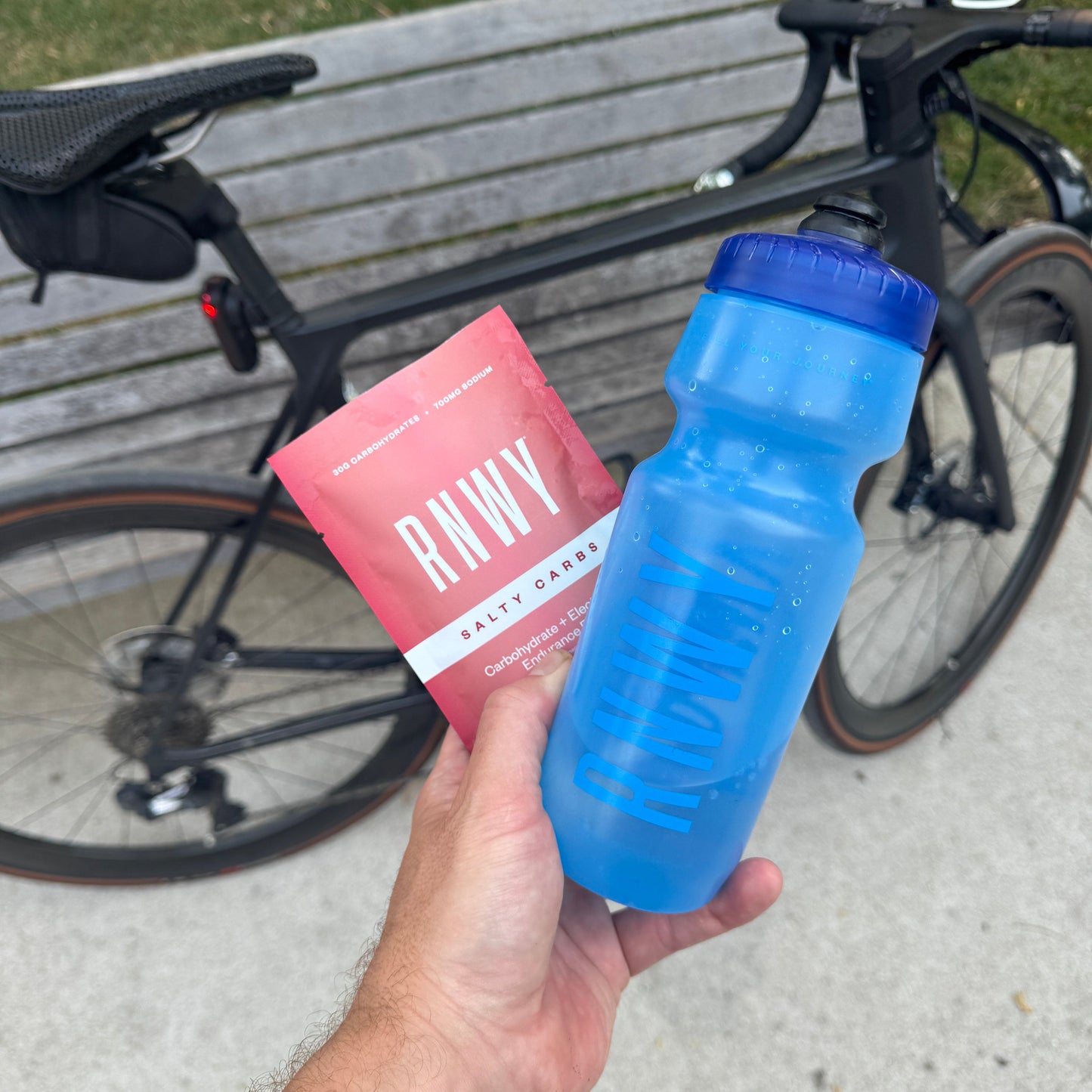 Hand holding a blue water bottle and pink packet with 'RNWY' branding in front of a bicycle.