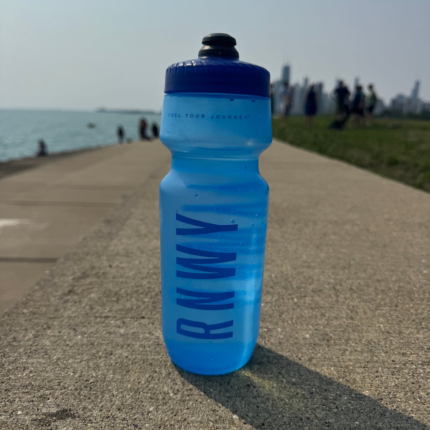 Blue water bottle with 'RNW' branding on a concrete surface near a waterfront with people in the background.